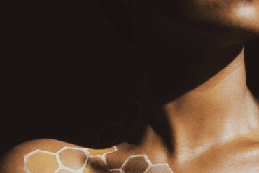 An cropped image of a Black woman with a honeycomb painted onto her body. Her eyes are closed and sunlight is streaming onto her face.