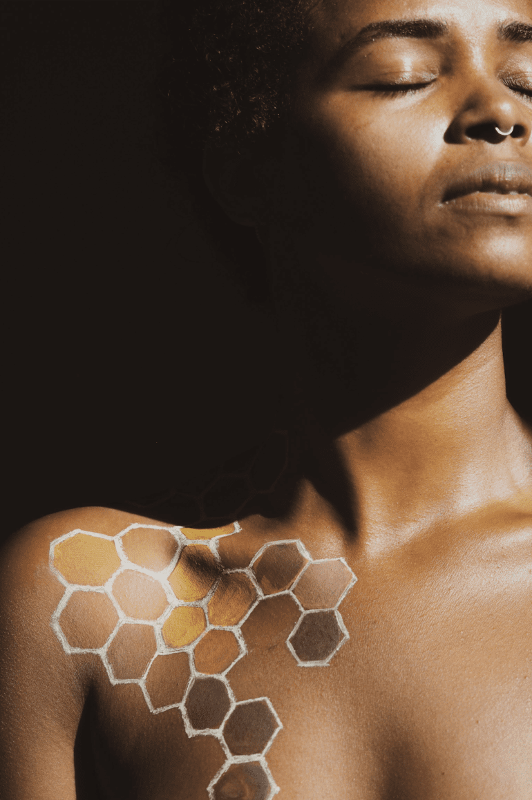 An cropped image of a Black woman with a honeycomb painted onto her body. Her eyes are closed and sunlight is streaming onto her face.